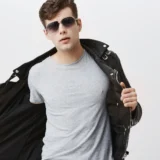 Joven con gafas de sol y chaqueta de cuero negra, luciendo una camiseta gris, posando con actitud segura frente a un fondo blanco.