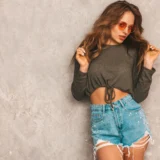 Mujer con blusa de manga larga gris y pantalones cortos de mezclilla decorados, posando frente a una pared gris, luciendo gafas de sol naranjas y mostrando un estilo moderno.