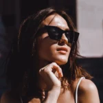 Mujer con gafas de sol negras, posando al aire libre con una expresión pensativa, cabello suelto y luz natural que resalta su piel. Fondo desenfocado con elementos arquitectónicos.
