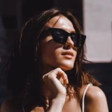 Mujer con gafas de sol negras, posando al aire libre con una expresión pensativa, cabello suelto y luz natural que resalta su piel. Fondo desenfocado con elementos arquitectónicos.