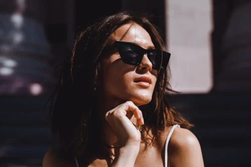 Mujer con gafas de sol negras, posando al aire libre con una expresión pensativa, cabello suelto y luz natural que resalta su piel. Fondo desenfocado con elementos arquitectónicos.