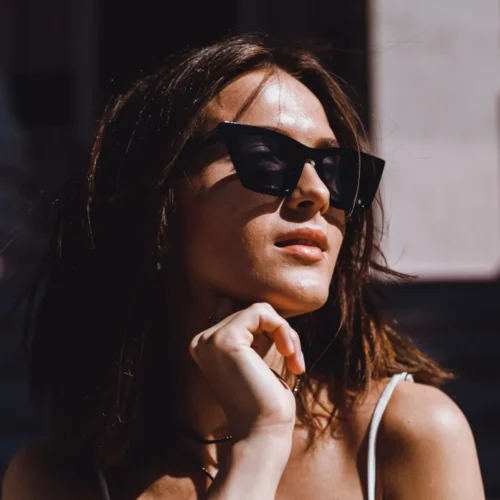 Mujer con gafas de sol negras, posando al aire libre con una expresión pensativa, cabello suelto y luz natural que resalta su piel. Fondo desenfocado con elementos arquitectónicos.